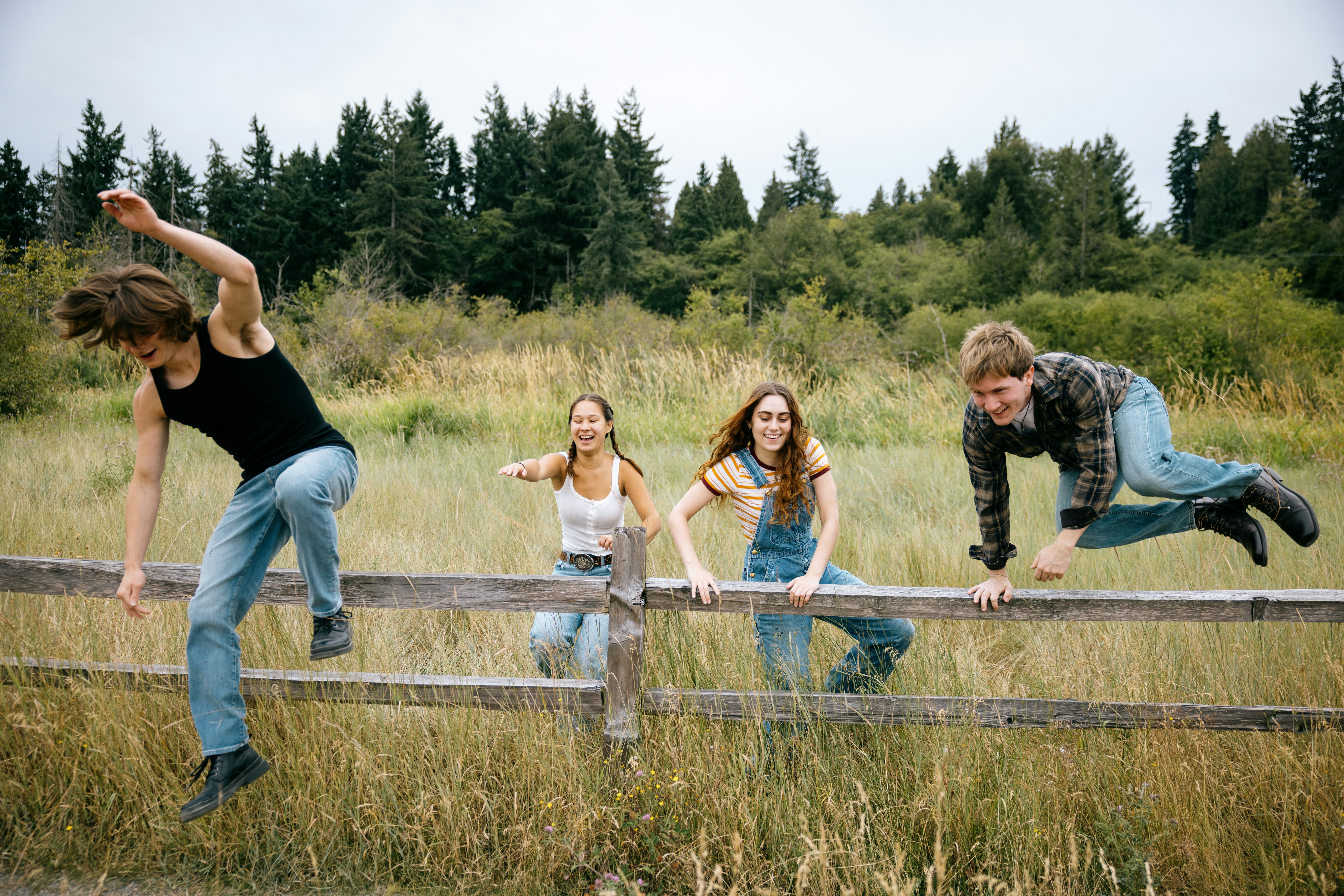 Four friends climbing over a wooden fence together in a grassy field, laughing and helping each other outdoors.
